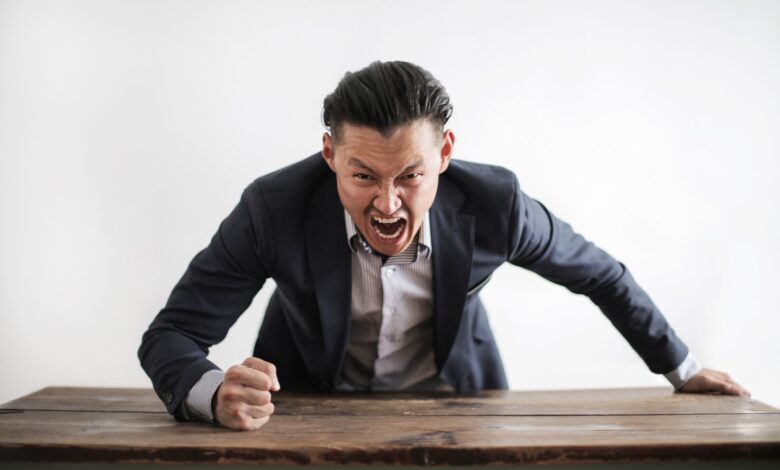 Expressive angry businessman in formal suit looking at camera and screaming with madness while hitting desk with fist