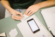 Close-up of student writing notes with a phone displaying diagrams. Concept of study aids and education.