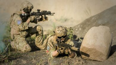 Two soldiers in camouflage uniforms practice tactical maneuvers in a desert environment, exhibiting teamwork and precision.