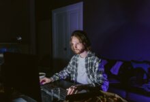 A man wearing headphones plays video games on a computer in a dark room, illuminated by screen light.