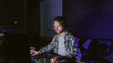 A man wearing headphones plays video games on a computer in a dark room, illuminated by screen light.