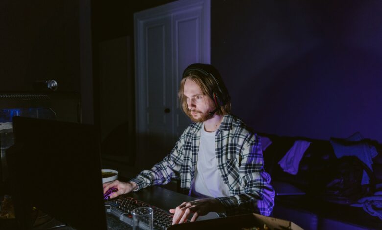 A man wearing headphones plays video games on a computer in a dark room, illuminated by screen light.