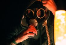 A dramatic image of a hooded individual wearing a gas mask illuminated by warm light, creating a mysterious atmosphere.