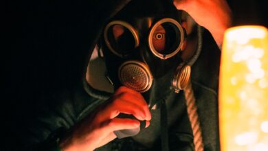 A dramatic image of a hooded individual wearing a gas mask illuminated by warm light, creating a mysterious atmosphere.