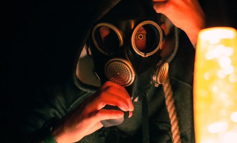 A dramatic image of a hooded individual wearing a gas mask illuminated by warm light, creating a mysterious atmosphere.