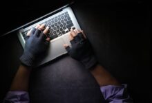 Hacker in fingerless gloves typing on laptop keyboard from above in a dark setting.