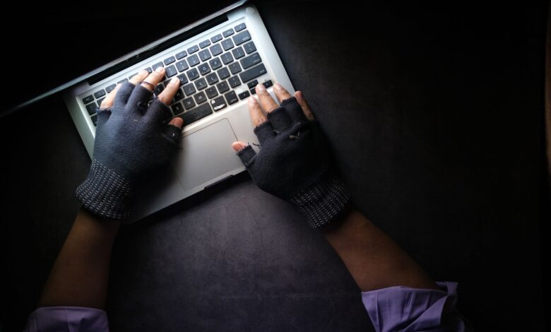 Hacker in fingerless gloves typing on laptop keyboard from above in a dark setting.