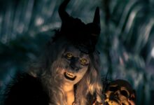 A costumed figure with horns and yellow eyes holds a skull in a dark forest setting, creating a spooky vibe.