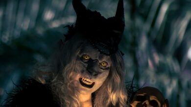 A costumed figure with horns and yellow eyes holds a skull in a dark forest setting, creating a spooky vibe.