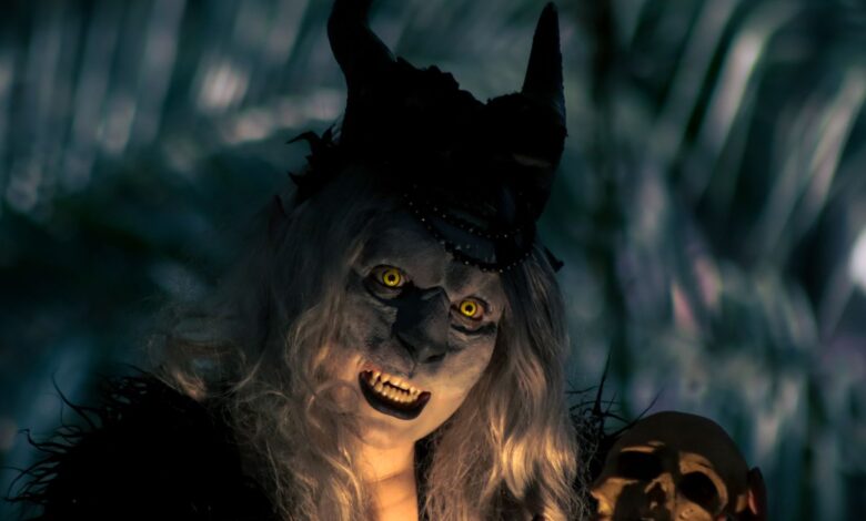 A costumed figure with horns and yellow eyes holds a skull in a dark forest setting, creating a spooky vibe.