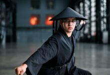 Martial artist wearing a black kimono and rice hat performing a striking pose indoors.