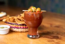 Chilled Mexican michelada garnished with olives and served alongside nachos on a wooden table.