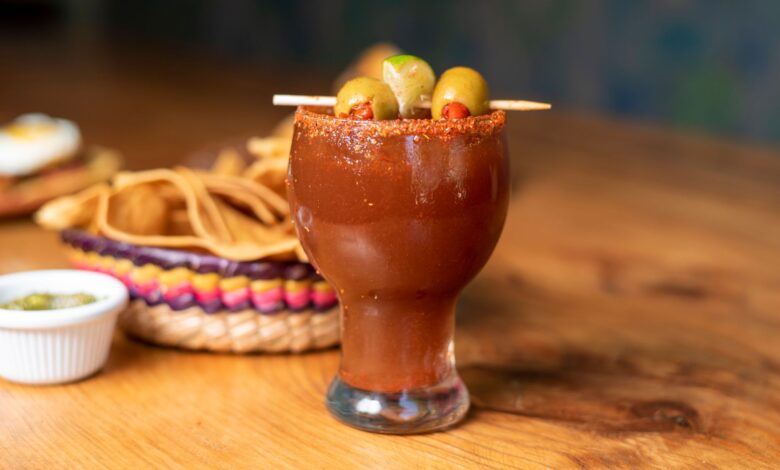 Chilled Mexican michelada garnished with olives and served alongside nachos on a wooden table.