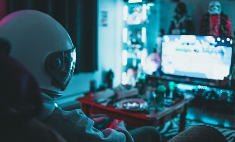 Side view of unrecognizable person in virtual reality helmet sitting on sofa and playing with gamepad in dark room