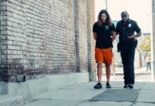 Police officer walking with handcuffed man along urban street with brick wall.