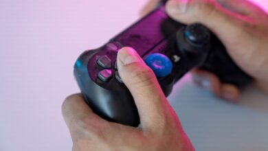 Close-up shot of hands gripping a black gaming controller with a blue thumbstick cover under neon lighting.