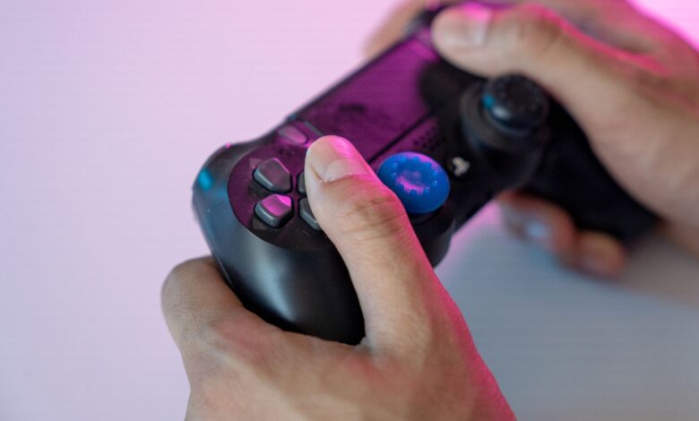 Close-up shot of hands gripping a black gaming controller with a blue thumbstick cover under neon lighting.
