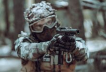 A focused soldier in tactical gear aiming a handgun in a forest setting.