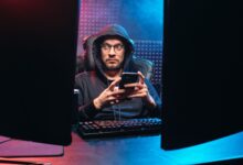A man in a hoodie using a smartphone, surrounded by tech gear in a dimly lit room.