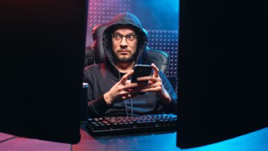 A man in a hoodie using a smartphone, surrounded by tech gear in a dimly lit room.