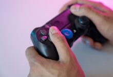 Close-up shot of hands gripping a black gaming controller with a blue thumbstick cover under neon lighting.