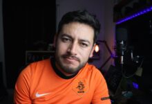 Close-up portrait of a man in an orange jersey next to a microphone in a cozy indoor studio.