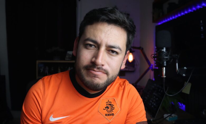 Close-up portrait of a man in an orange jersey next to a microphone in a cozy indoor studio.