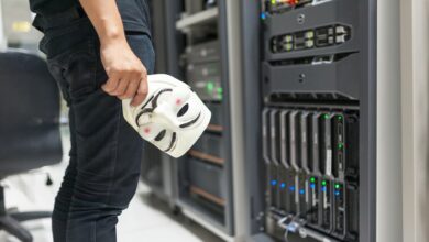 Person holding anonymous mask near servers, hinting at cybersecurity and hacking themes.