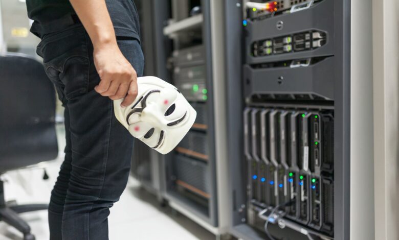 Person holding anonymous mask near servers, hinting at cybersecurity and hacking themes.