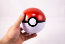 Close-up of a human hand holding a red Pokeball toy against a white background.