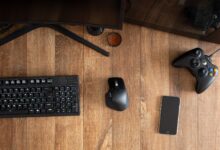 Top view of black gamepad with mouse and keyboard on wooden desk with monitor in apartment in daytime