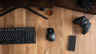 Top view of black gamepad with mouse and keyboard on wooden desk with monitor in apartment in daytime