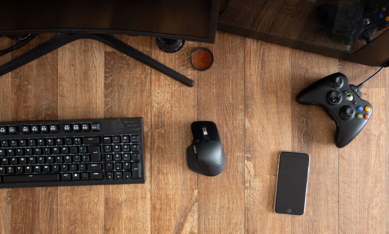 Top view of black gamepad with mouse and keyboard on wooden desk with monitor in apartment in daytime