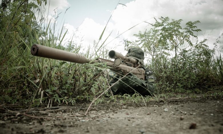 A camouflaged sniper with rifle hidden in grasslands during daytime surveillance.