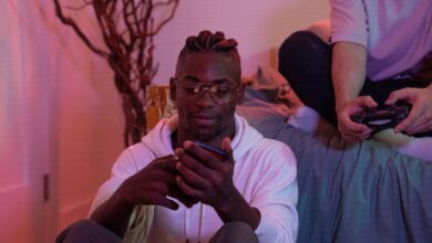 Two friends enjoying a gaming session at home, one using a mobile phone and the other a game controller.
