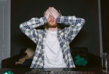 Focused male gamer in plaid shirt, showing stress with hands on face, indoors.