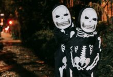 Anonymous kids in skeleton costumes and masks embracing while standing on street in evening on blurred background during Halloween celebration