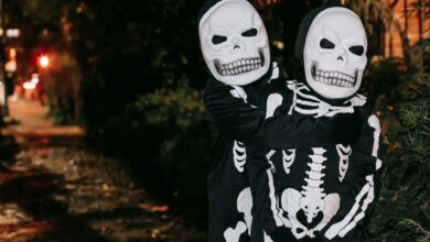 Anonymous kids in skeleton costumes and masks embracing while standing on street in evening on blurred background during Halloween celebration
