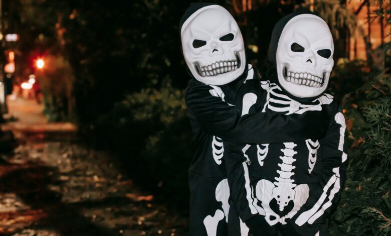 Anonymous kids in skeleton costumes and masks embracing while standing on street in evening on blurred background during Halloween celebration
