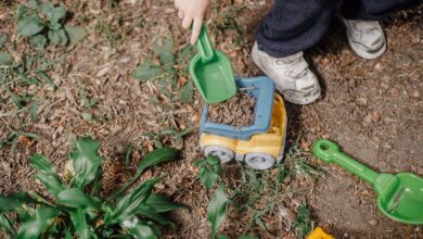Top view of anonymous playful child picking soil into plastic toy car while playing on ground with green plants on street