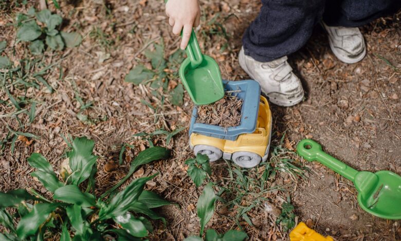 Top view of anonymous playful child picking soil into plastic toy car while playing on ground with green plants on street