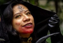 Close-up of a woman in vampire cosplay, featuring a rose tattoo and yellow contact lenses, outdoors.