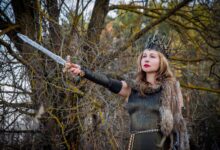 A fierce warrior queen in medieval costume holding a sword against a forest backdrop.