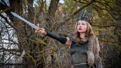 A fierce warrior queen in medieval costume holding a sword against a forest backdrop.