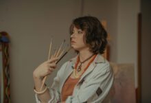 A young female artist stands in her studio, holding paintbrushes with a serene expression.