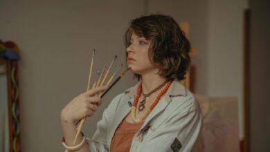 A young female artist stands in her studio, holding paintbrushes with a serene expression.