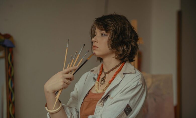 A young female artist stands in her studio, holding paintbrushes with a serene expression.