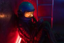 A woman in a space suit sits alone in a dimly lit spacecraft with red and blue lights.