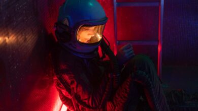 A woman in a space suit sits alone in a dimly lit spacecraft with red and blue lights.