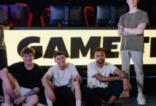 Four young men posing confidently in an eSports arena with gaming setups in the background.